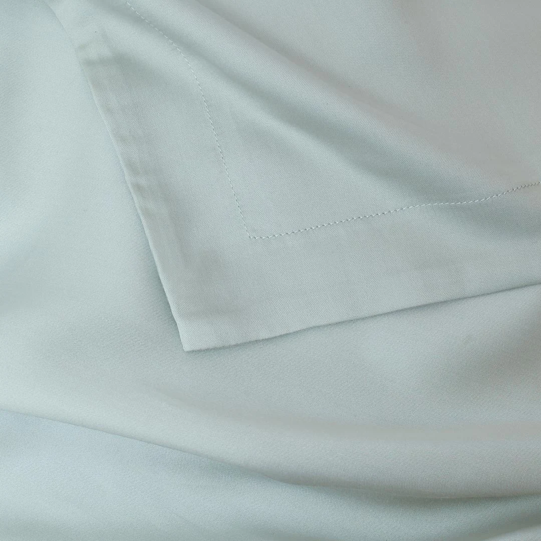 Close-up van een lichtblauwe kussensloop. De stof is glad en heeft een subtiele glans. De hoes is netjes afgewerkt met een stiksel.