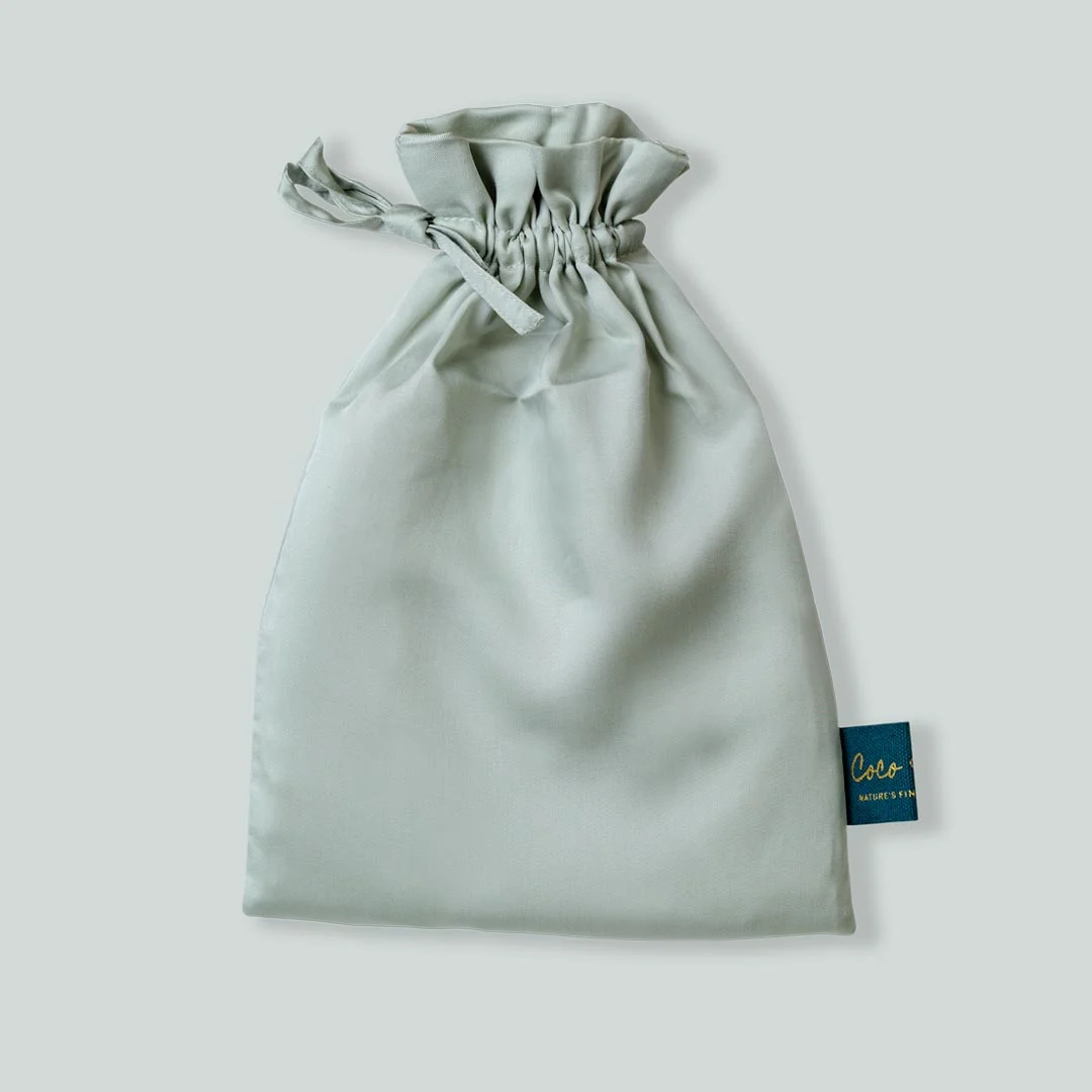 Zachtgrijze Tencel kussensloop verpakt in een satijnen zakje met trekkoord, label rechtsonder. Studiofotografie, zachte belichting.