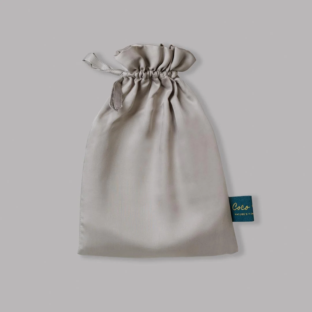Grijze satijnen zak met trekkoord, zachte stof. Label 'Coco Nature & Kind' zichtbaar. Neutrale achtergrond. Ideaal voor kussensloop.