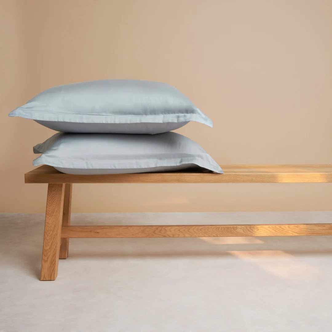 Twee Kussensloop 60x70 Grijs Comfortabele touch kussenslopen liggen gestapeld op een houten bank tegen een beige muur en een lichtgekleurde vloer.