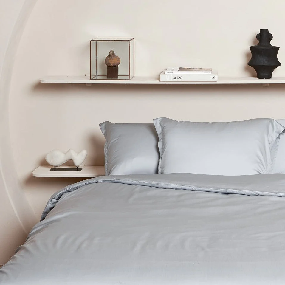 Een netjes opgemaakt bed met de Kussensloop 60x70 Grijs voor een comfortabele touch. Tegen een beige muur zorgt een zwevende plank met abstracte beelden en boeken voor een minimalistische, moderne inrichting boven het bed.