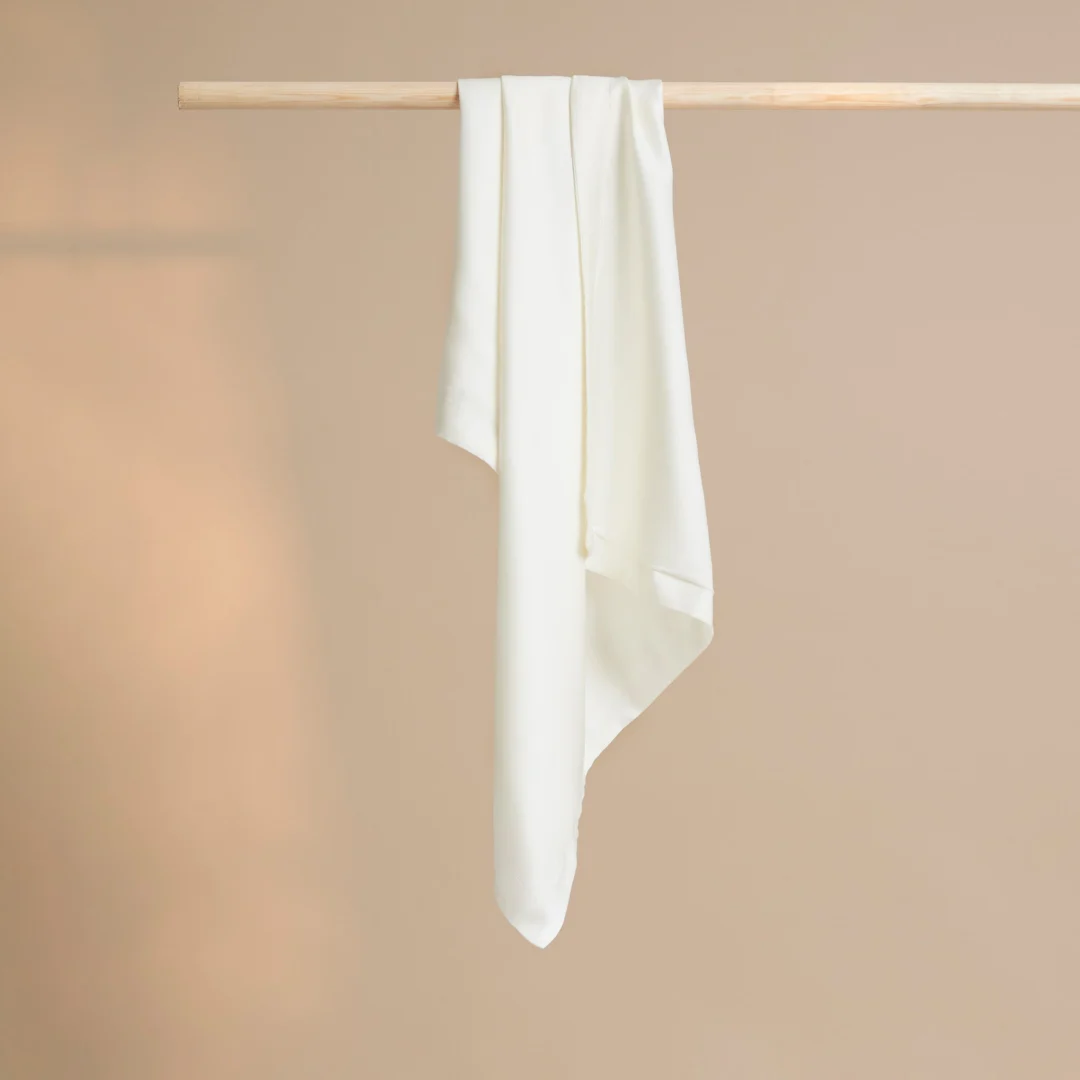 De Kussensloop 60x70 Risse uitstraling, een lange witte kussensloop, hangt gedrapeerd over een eenvoudige houten roede tegen een effen beige achtergrond.