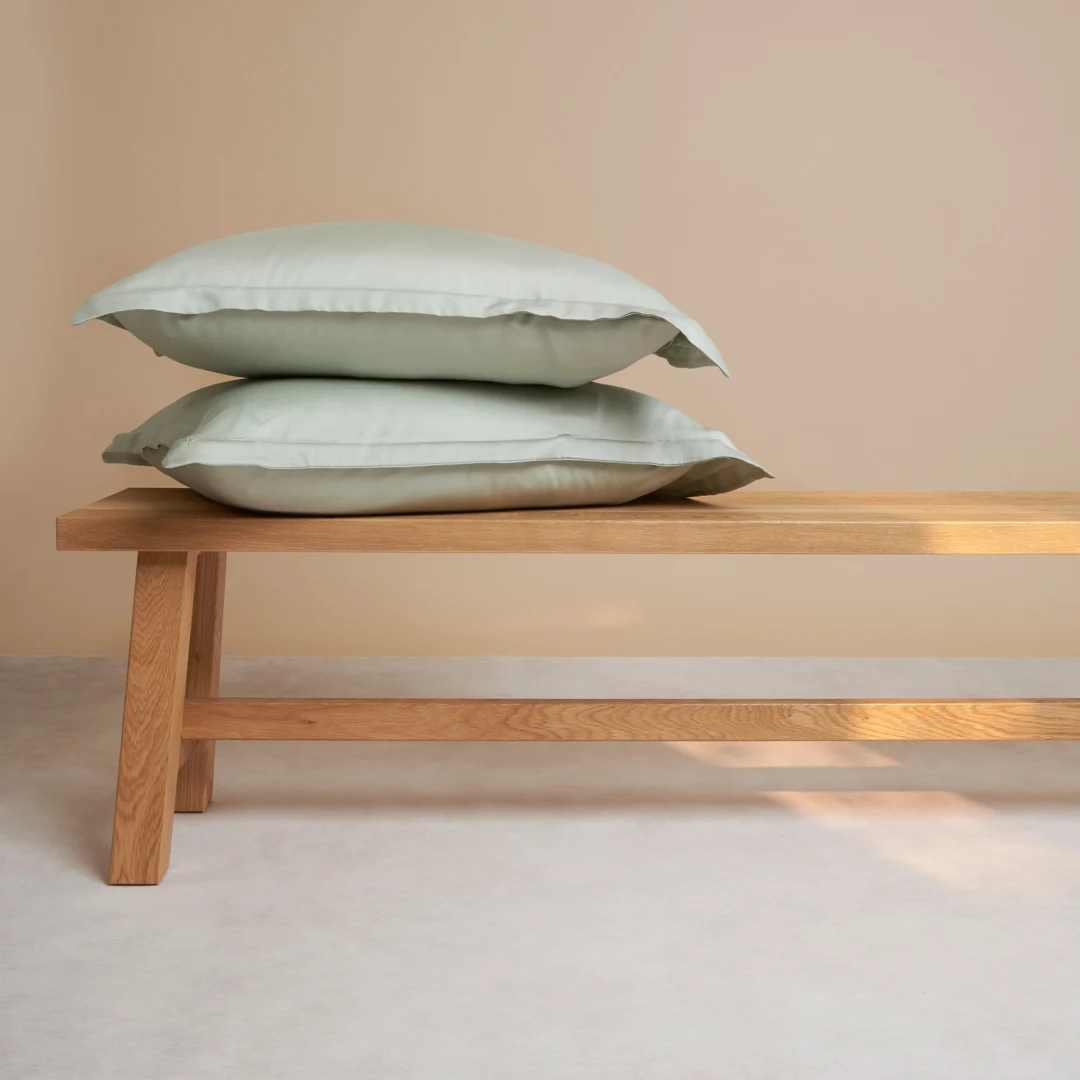 Een Dekbedovertrek 60x70 Grijs Comfortabele touch is opgevouwen uitgestald op een eenvoudige houten bank, tegen een beige muur en een lichtgekleurde vloer.