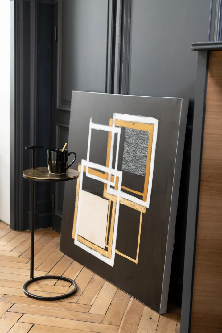 Een modern abstract schilderij met geometrische vormen in wit, goud en grijs leunt tegen een donkergrijze muur op een houten vloer, naast de Sidetable Toni Aluminium/IJzer Goud/Zwart, die een zwarte mok met een gouden lepel vasthoudt.