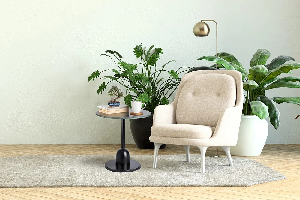 Naast de Bijzettafel Groen Rond Marmer staat een gezellige beige fauteuil met boeken, een plant en een mok. Grote groene kamerplanten en een gouden staande lamp accentueren de luchtige woonkamer met een grijs vloerkleed en houten vloeren.
