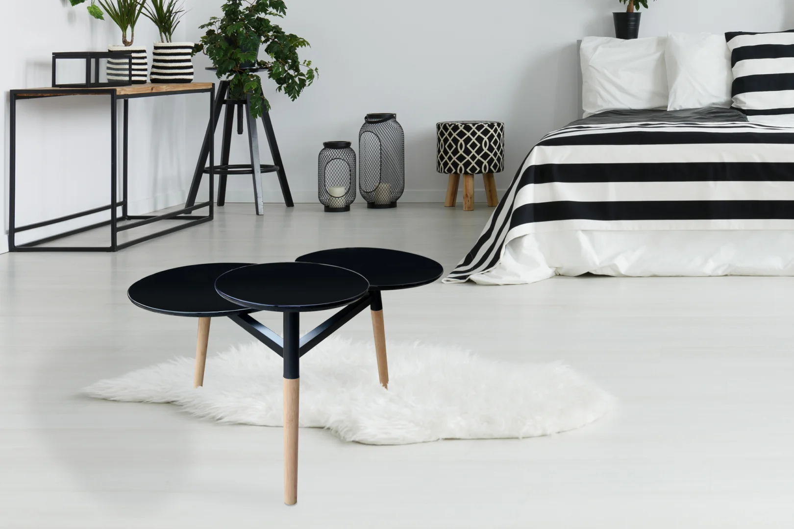 Een moderne slaapkamer heeft een zwart-wit gestreepte sprei, een Bijzettafel Zwart 30 CM Rond Bamboe met opbergvak, een pluizig wit vloerkleed, planten, decoratieve lantaarns en een heldere minimalistische inrichting.