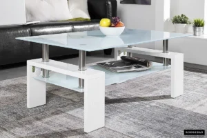 De Salontafel Wit Design Modern is een moderne glazen salontafel met een wit onderstel, afgebeeld met een schaal met divers fruit erop en tijdschriften op de onderste plank, op een grijs vloerkleed in een hedendaagse woonkamer.