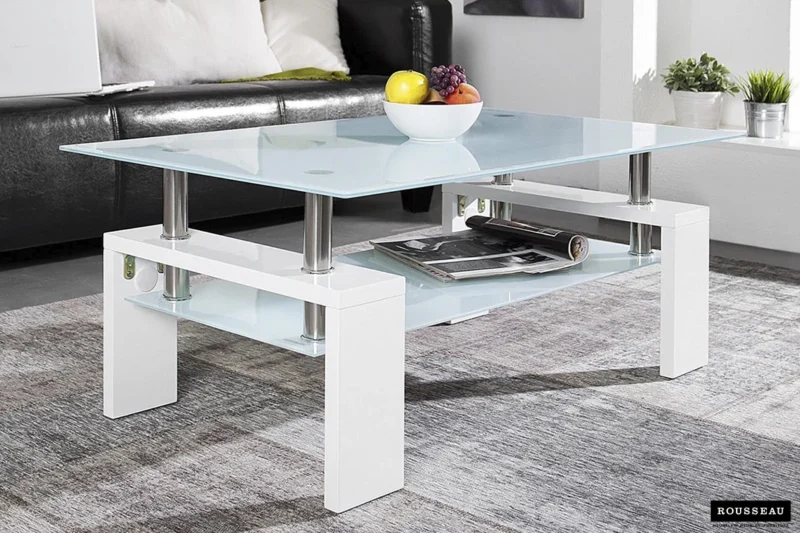 De Salontafel Wit Design Modern is een moderne glazen salontafel met een wit onderstel, afgebeeld met een schaal met divers fruit erop en tijdschriften op de onderste plank, op een grijs vloerkleed in een hedendaagse woonkamer.