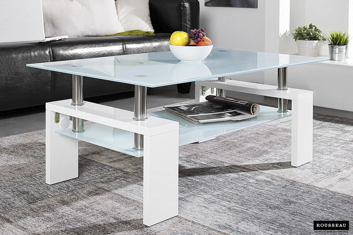 De Salontafel Wit Design Modern is een moderne glazen salontafel met een wit onderstel, afgebeeld met een schaal met divers fruit erop en tijdschriften op de onderste plank, op een grijs vloerkleed in een hedendaagse woonkamer.