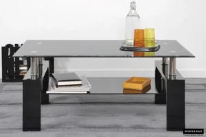 De Salontafel Zwart Tijdloos Design, met zwarte poten, heeft een glazen blad met een heldere fles, een sinaasappel en een geel glas op een dienblad. Boeken en een gele doos staan op de onderste plank en de tafel staat op een grijs vloerkleed.