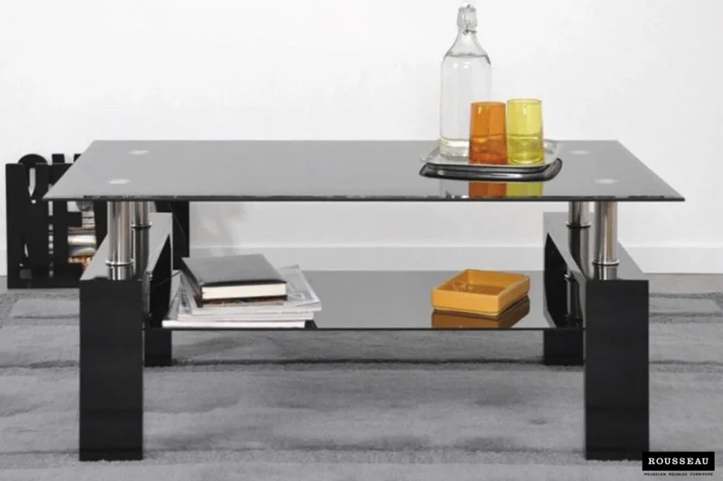 De Salontafel Zwart Tijdloos Design, met zwarte poten, heeft een glazen blad met een heldere fles, een sinaasappel en een geel glas op een dienblad. Boeken en een gele doos staan op de onderste plank en de tafel staat op een grijs vloerkleed.