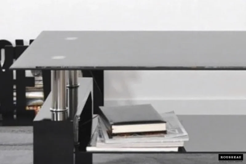 De Salontafel Zwart Tijdloos Design is een moderne zwarte salontafel met metalen cilindrische steunen en een minimalistische uitstraling, getoond met boeken en tijdschriften op de onderste plank tegen een witte muur.