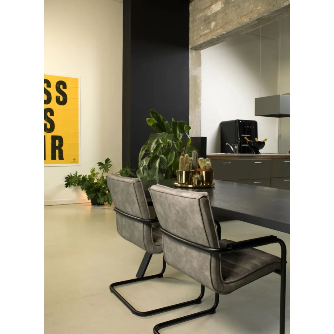 Moderne eethoek met twee Werkleven Haven Eetkamerstoel - Vegan Leer Grijs stoelen aan een zwarte tafel, planten in potten, gele muurkunst op de achtergrond en een glimp van de keuken aan de rechterkant. Minimalistische industriële stijl.