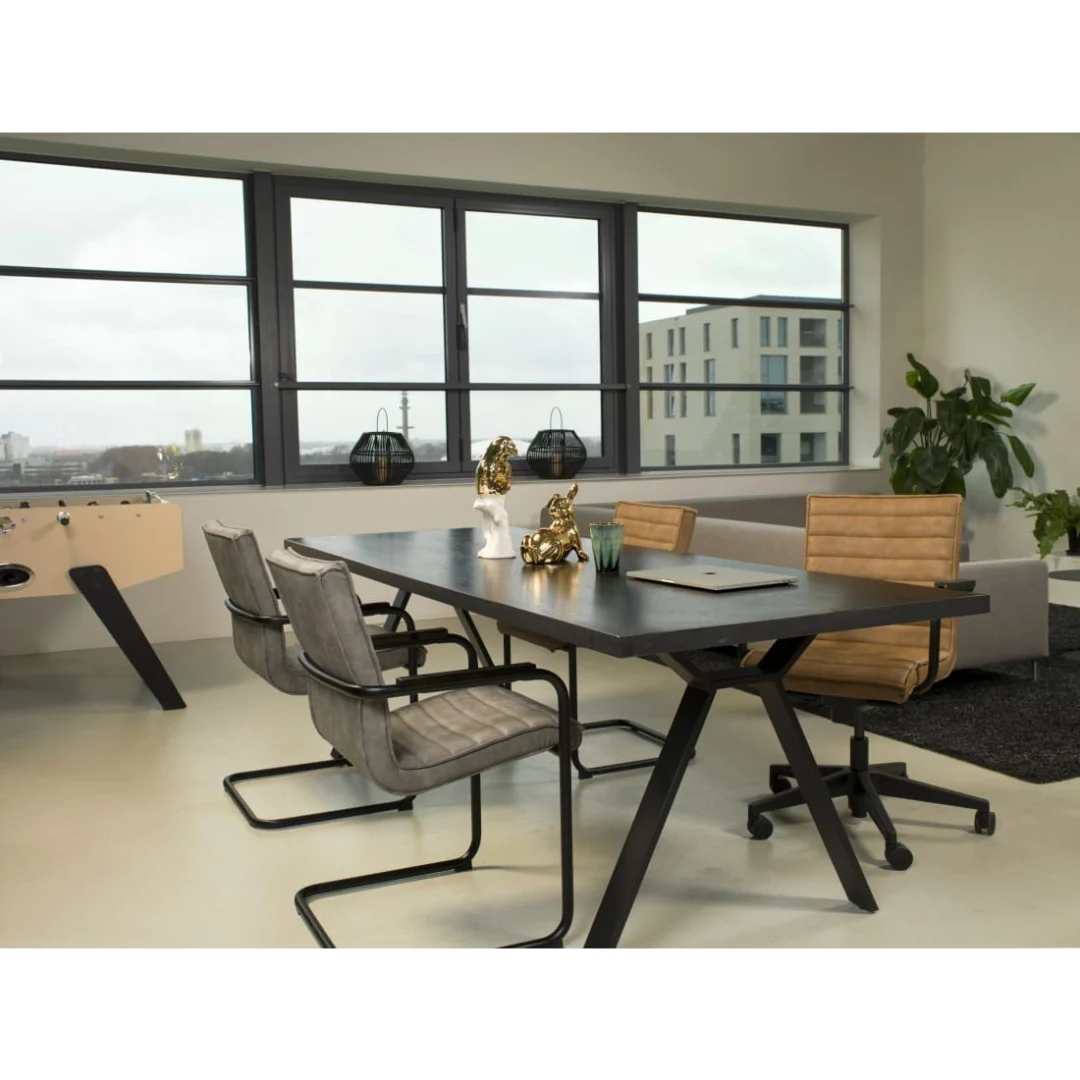 Een modern kantoor met een groot zwart bureau, een laptop, decoratieve voorwerpen, een bank, een plant en grote ramen met uitzicht op de stad. Vier Werkleven Haven Eetkamerstoel - Vegan Leer Grijs stoelen omringen het bureau terwijl natuurlijk licht de kamer vult.