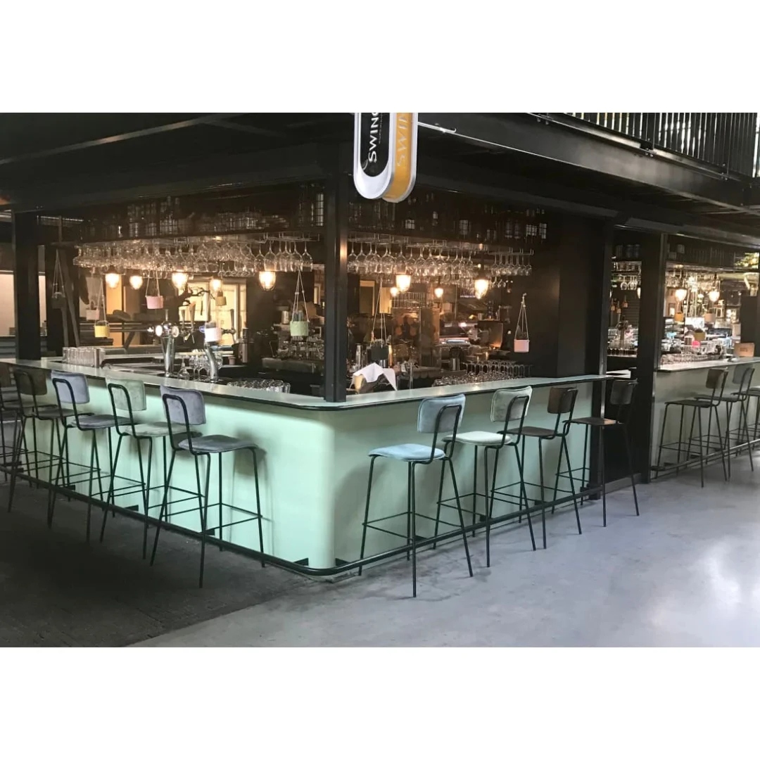 Een moderne binnenbar met Werkwoonkruk Hoog Bo - Fluweel Aardeleger hoge barkrukken, een lichtgroene toonbank, hangend glaswerk, zachte verlichting en spiegelende planken met flessen achter de bar voor een gezellige sfeer.