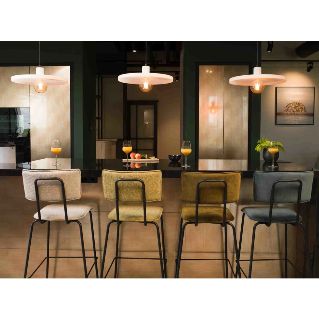 Vier Werkwoonkruk Hoog Bo - Fluweel Aarde Langs een keukeneiland staan groenblauwe barkrukken met daarop drie glazen sinaasappelsap, een plantje en fruit. Erboven hangen hanglampen die een modern tintje aan het tafereel geven.