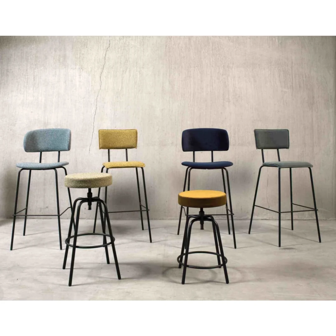 Een set van zes Werkwoonkruk Hoog Bo Society barkrukken met zwarte metalen poten en Boucle Mosterd bekleding, plus andere kleuren (blauw, geel, grijs, groen), staan opgesteld op een betonnen vloer tegen een grijze muur met structuur.