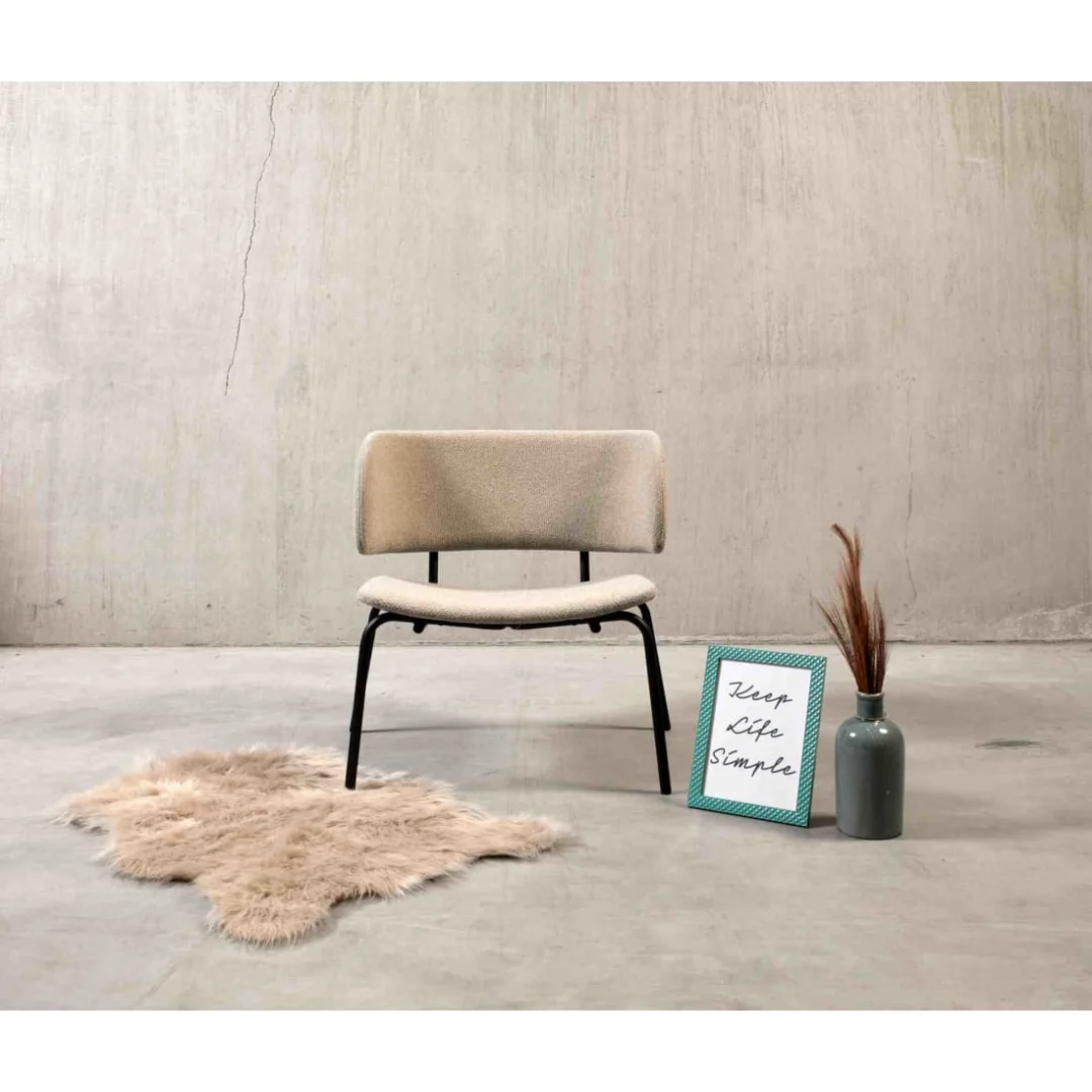 Een minimalistische kamer met de Werkwoonkamer Bo Lounge Fauteuil - Society Boucle Shitake, een klein harig vloerkleed, een vaas met gedroogd gras en een ingelijst bord "Keep Life Simple" tegen lichte betonnen muren en vloer.