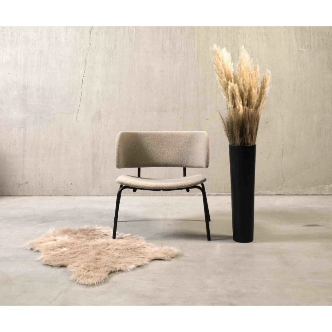 De Werkwoonkamer Bo Lounge Fauteuil - Society Boucle Shitake in beige met zwarte poten staat op een betonnen vloer naast een hoge zwarte vaas met pampagras en een donzig beige vloerkleed, tegen een effen, lichtgekleurde muur.