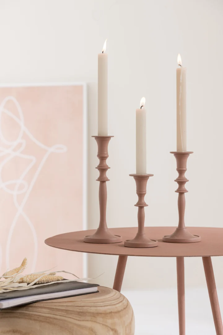 Drie brandende witte kaarsen in roze kandelaars staan op de Bijzettafel Roze 56 x 56 CM Rond Ijzer, met abstracte kunst en een stapel boeken met gedroogde bloemen op de achtergrond. Het tafereel heeft een minimalistisch, modern decor.