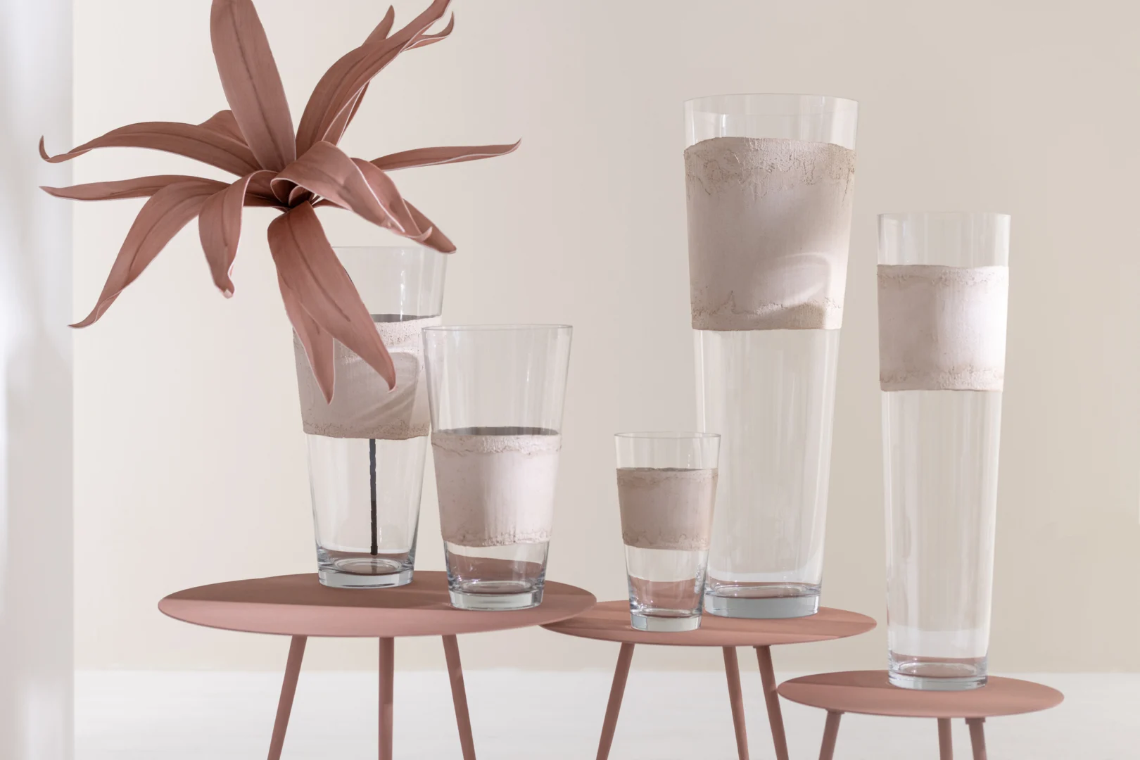 Vijf heldere glazen met gestructureerde beige banden en een sculpturale bruine plant zijn gerangschikt op drie Bijzettafel Roze 56 x 56 CM Rond Ijzer, waardoor een minimalistische en moderne display ontstaat tegen een neutrale achtergrond.
