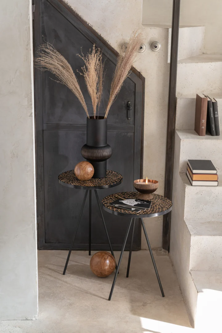 Een Bijzettafel Zwart, Bruin (38 x 45 CM) met metalen afwerking staat bij een beige muur, versierd met een zwarte vaas met gedroogd gras, een houten bol en een brandende kaars. Er liggen boeken op de nabijgelegen stenen val.