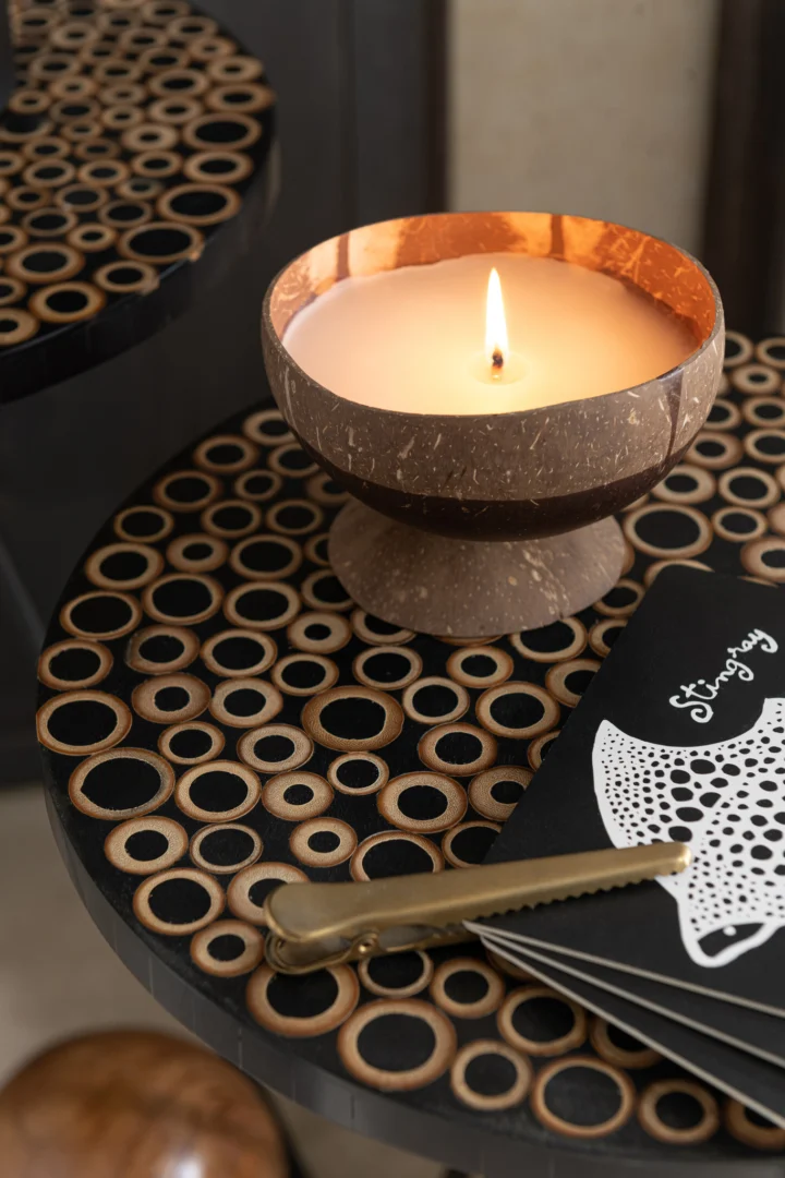 Een brandende kaars in een kokosnootschaal rust op de Bijzettafel Zwart, Bruin (38 x 45 CM, metaal), naast een zwart notitieboekje met wit kanten detail en een gouden lontsnijder.