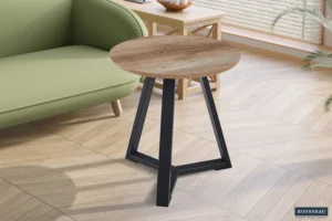De bijzettafel 'Paso 2' Decor Old Oak, met een klein rond houten blad en zwarte metalen poten, staat naast een groene bank in een lichte kamer met houten vloer, moderne inrichting en zonlicht dat door de jaloezieën naar binnen schijnt.
