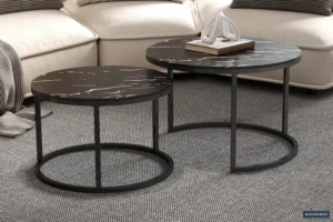 De Salontafel Zwart, Roest Marmer - twee ronde zwarte tafels met wit gemarmerd design en metalen frames - staan op een grijs gestructureerd tapijt voor een beige bank, met boeken en decor op de grotere tafel.