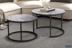 De Salontafel Grijs 35 CM Steen, een ronde grijze salontafel met een metalen frame, staat op een grijs vloerkleed met textuur voor een beige sofa met gestapelde boeken en een decoratieve sculptuur.