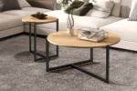 Twee Salontafel Klassiek Design Tijdloos tafels met zwarte metalen poten staan op een grijs tapijt in de woonkamer. Op de ene staat een glazen vaas met groen, op de andere een schaal met dennenappels. Een beige sofa verschijnt op de achtergrond.