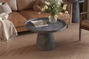 De Salontafel Donkergrijs, Roest 80 CM met een gestructureerde afwerking staat op een lichthouten visgraatvloer, met daarop een vaas met witte tulpen en een tijdschrift. Op de achtergrond staat een bruine bank gedrapeerd met een kleed.
