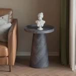 Een Salontafel Donkergrijs, Roest 45 CM met een donkere afwerking houdt een witte klassieke buste vast naast een tan leren bank met grijze en beige kussens, dit alles op een houten vloer met visgraatmotief.