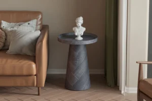 Een Salontafel Donkergrijs, Roest 45 CM met een donkere afwerking houdt een witte klassieke buste vast naast een tan leren bank met grijze en beige kussens, dit alles op een houten vloer met visgraatmotief.