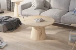De Salontafel 80 CM Rond, een ronde salontafel van licht hout, staat op een bijpassende vloer in een moderne woonkamer met een grijze bank, kussens met patroon en gezellige plaids. Natuurlijk licht stroomt naar binnen vanuit het linkerraam.