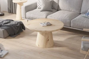 De Salontafel 80 CM Rond, een ronde salontafel van licht hout, staat op een bijpassende vloer in een moderne woonkamer met een grijze bank, kussens met patroon en gezellige plaids. Natuurlijk licht stroomt naar binnen vanuit het linkerraam.