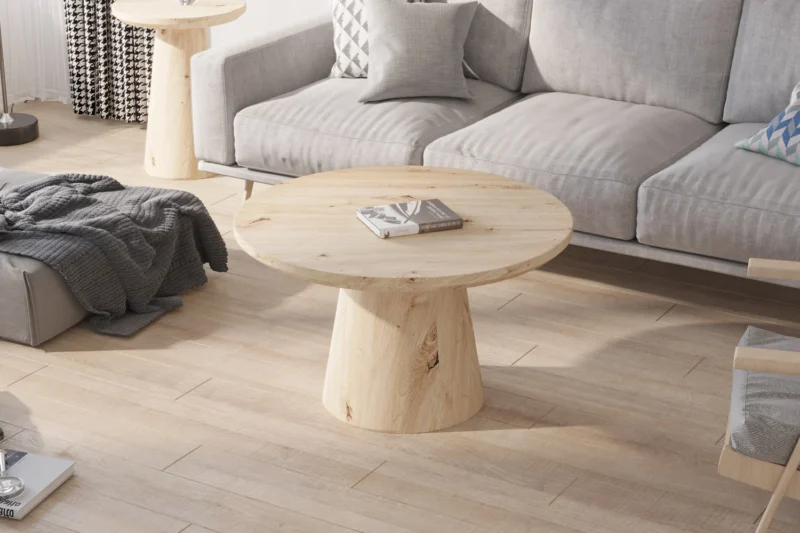 De Salontafel 80 CM Rond, een ronde salontafel van licht hout, staat op een bijpassende vloer in een moderne woonkamer met een grijze bank, kussens met patroon en gezellige plaids. Natuurlijk licht stroomt naar binnen vanuit het linkerraam.