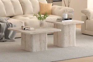 Twee Salontafel Beige Travertin vierkante salontafels met gecanneleerde voet staan op een gedessineerd tapijt voor een beige sofa. Op de ene staat een heldere vaas met groene stelen; op de andere staan gestapelde boeken en een wit decorstuk.