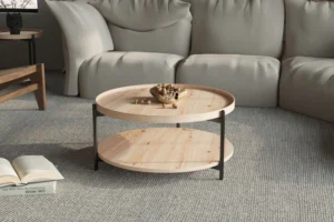 De Salontafel Roest 80 CM, een ronde houten salontafel met zwarte metalen poten, staat op een beige tapijt voor een lichtgekleurde sofa. Een schaal met noten staat op tafel en een open boek ligt op de vloer.