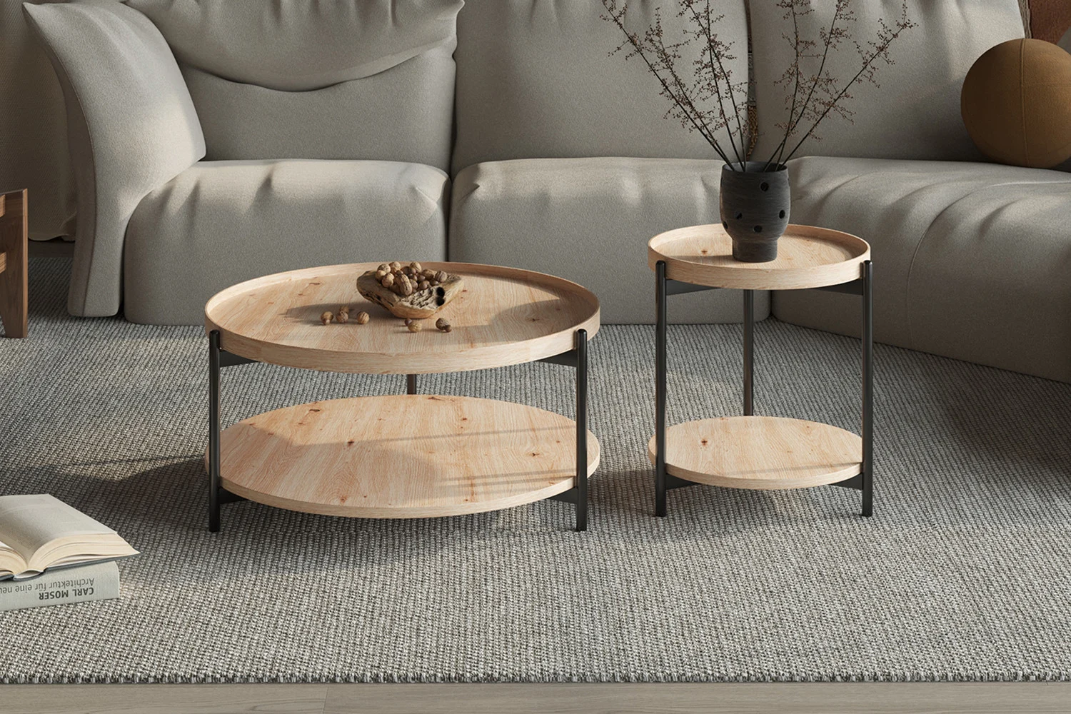 Een moderne woonkamer met een lichtgrijze bank en de Salontafel Roest 80 CM, een ronde houten salontafel met zwart metalen poten. Op de tafel staan een vaas met takken en een schaal met noten, allemaal geplaatst op een grijs vloerkleed met structuur.