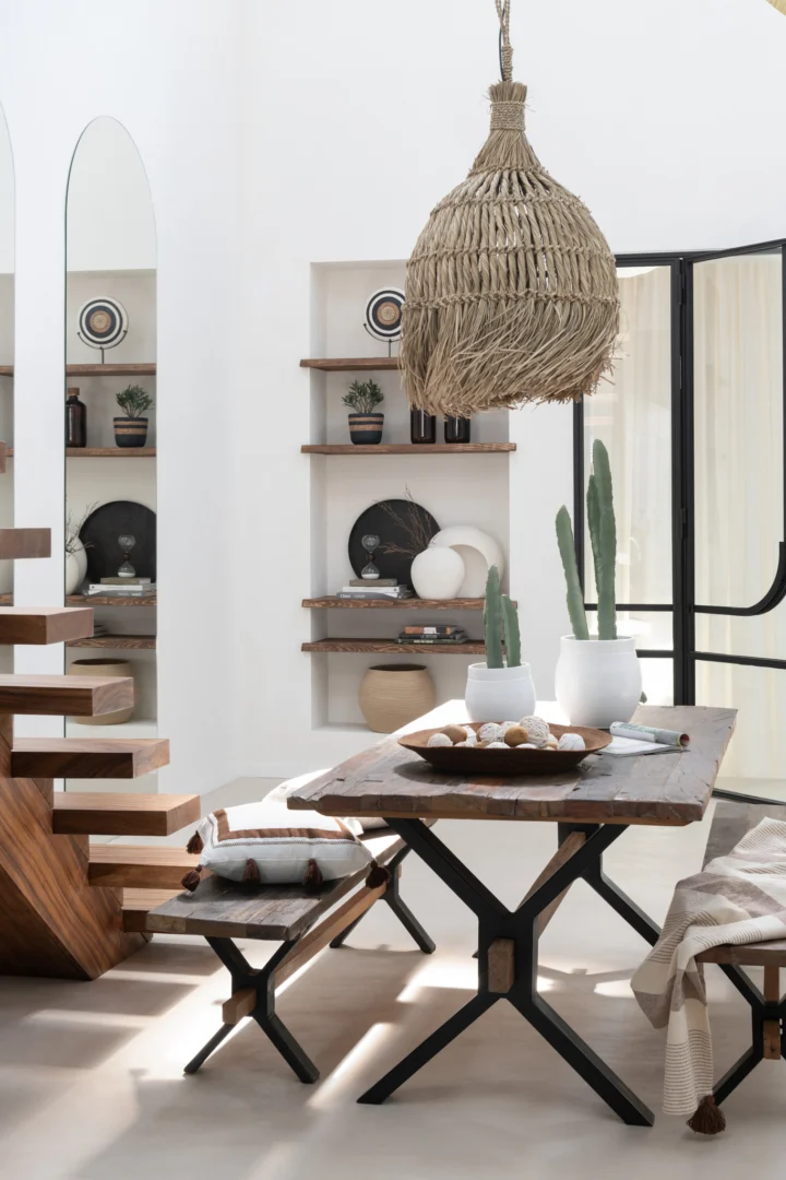 Een lichte, moderne eethoek is voorzien van de Tafel Vintage Hout Bruin met banken, cactussen in potten, zwevende decorplanken, een geweven hanglamp en een open trap met houten treden. De kamer is gevuld met natuurlijk licht.