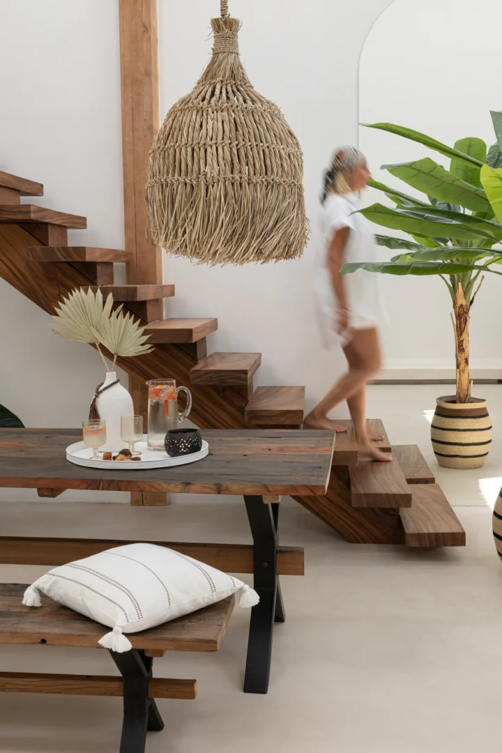 Een vage vrouw in een witte jurk loopt een houten trap af in een moderne, minimalistische kamer met de Tafel Vintage Hout Bruin, een houten bank, geweven hanglamp en grote potplanten.