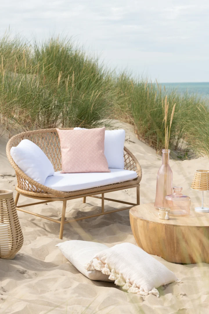 Een rieten loveseat met witte kussens en een roze kussen staat op zandduinen naast de Salontafel 32 CM Mangohout, waar roze glazen flessen en een lantaarn staan. Op de achtergrond zijn hoge grassen en de zee te zien.