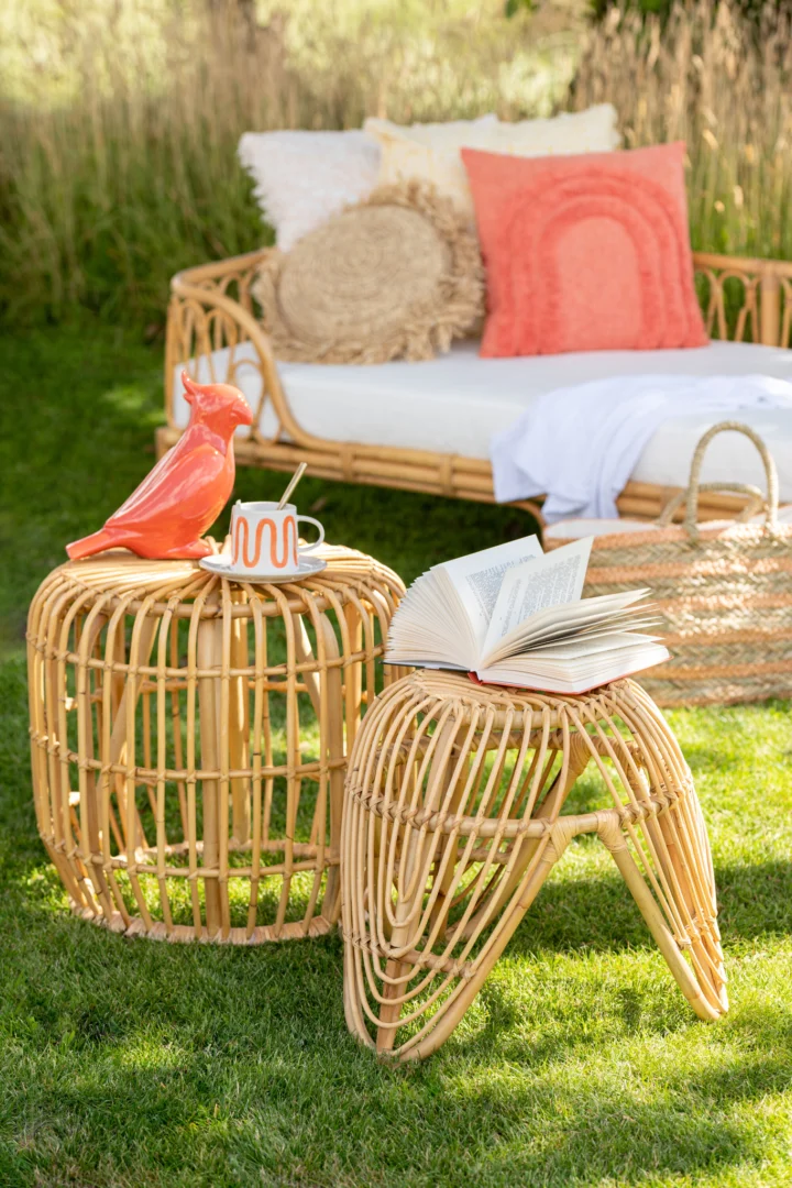 Een knusse buitenscène met een rotan daybed, koraalkleurige en beige kussens en twee Bijzettafel 45 x 35 CM Rotan bijzettafels - de ene met een vogelfiguurtje en mok, de andere met een open boek - op groen gras.