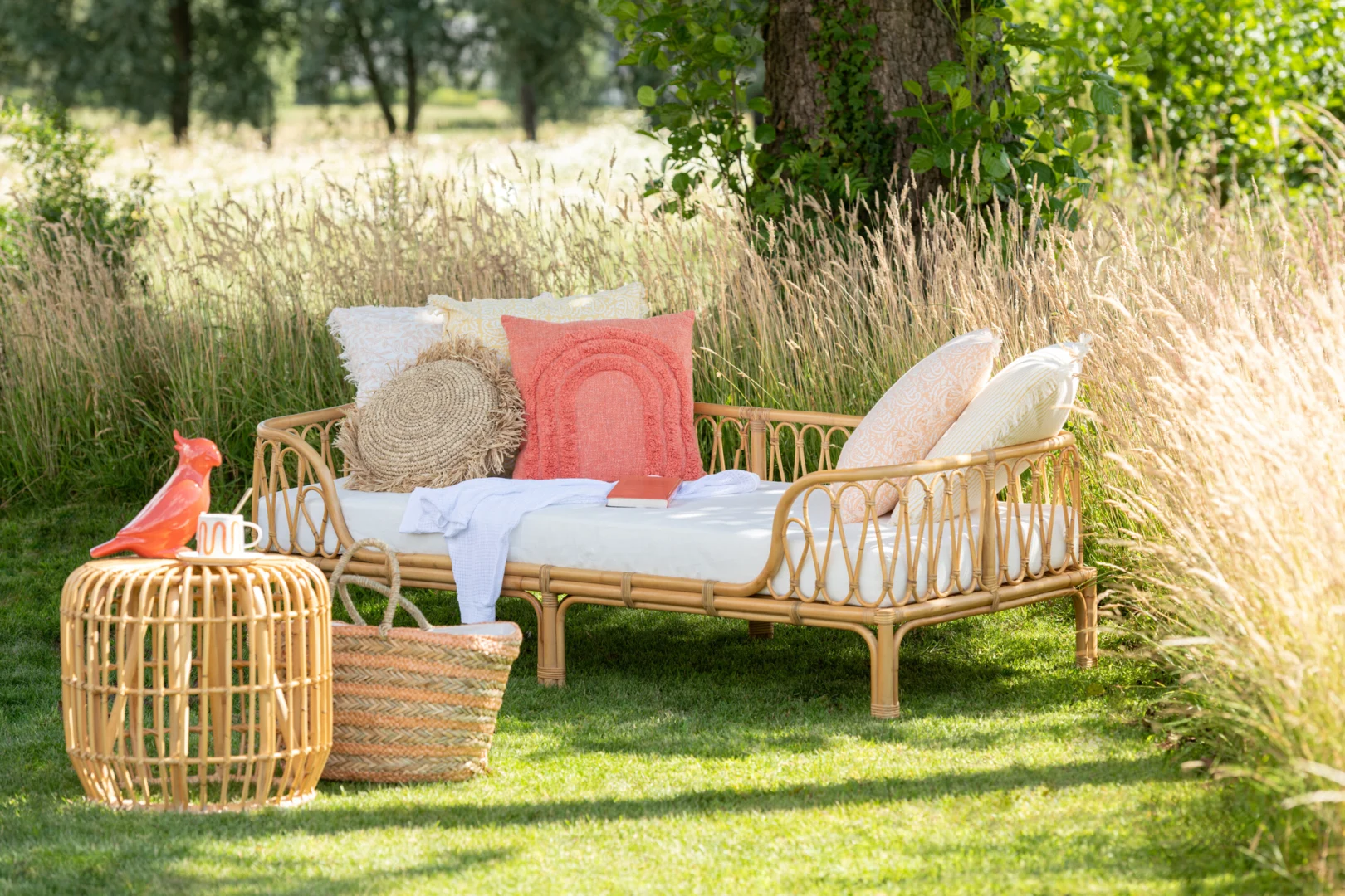 Een rotan daybed met kleurrijke kussens ligt op groen gras in een zonnige tuin, naast de Bijzettafel Rotan 45 x 35 CM met een koraalkleurig vogelornament en geweven mand, omringd door hoge grassen en bomen.