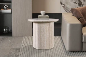 In een moderne woonkamer staat de Salontafel Beige 50 CM Rond Travertin - een geribbelde, ronde beige stenen bijzettafel met boeken en decor erop, naast een beige bank op een vloerkleed met structuur.