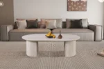 Een moderne woonkamer met een beige bank, neutrale kussens, een tapijt met structuur en de Salontafel Beige 120 CM Ovaal Travertin als salontafel met fruit en vazen. Abstracte kunst siert de muur achter de bank.