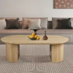 Een moderne woonkamer heeft een beige bank met neutrale kussens, een licht houten Salontafel 120 CM ovale salontafel met gebogen poten, met daarop een fruitschaal en een decoratieve vaas. Boven de bank hangt kunst met een patroon.