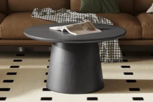 De Salontafel 80 CM Rond Design, een ronde zwarte houten salontafel met een open boek erop, staat op een crèmekleurig vloerkleed met zwarte vierkanten. Op de achtergrond staat een bruine bank gedrapeerd met een houndstooth deken.