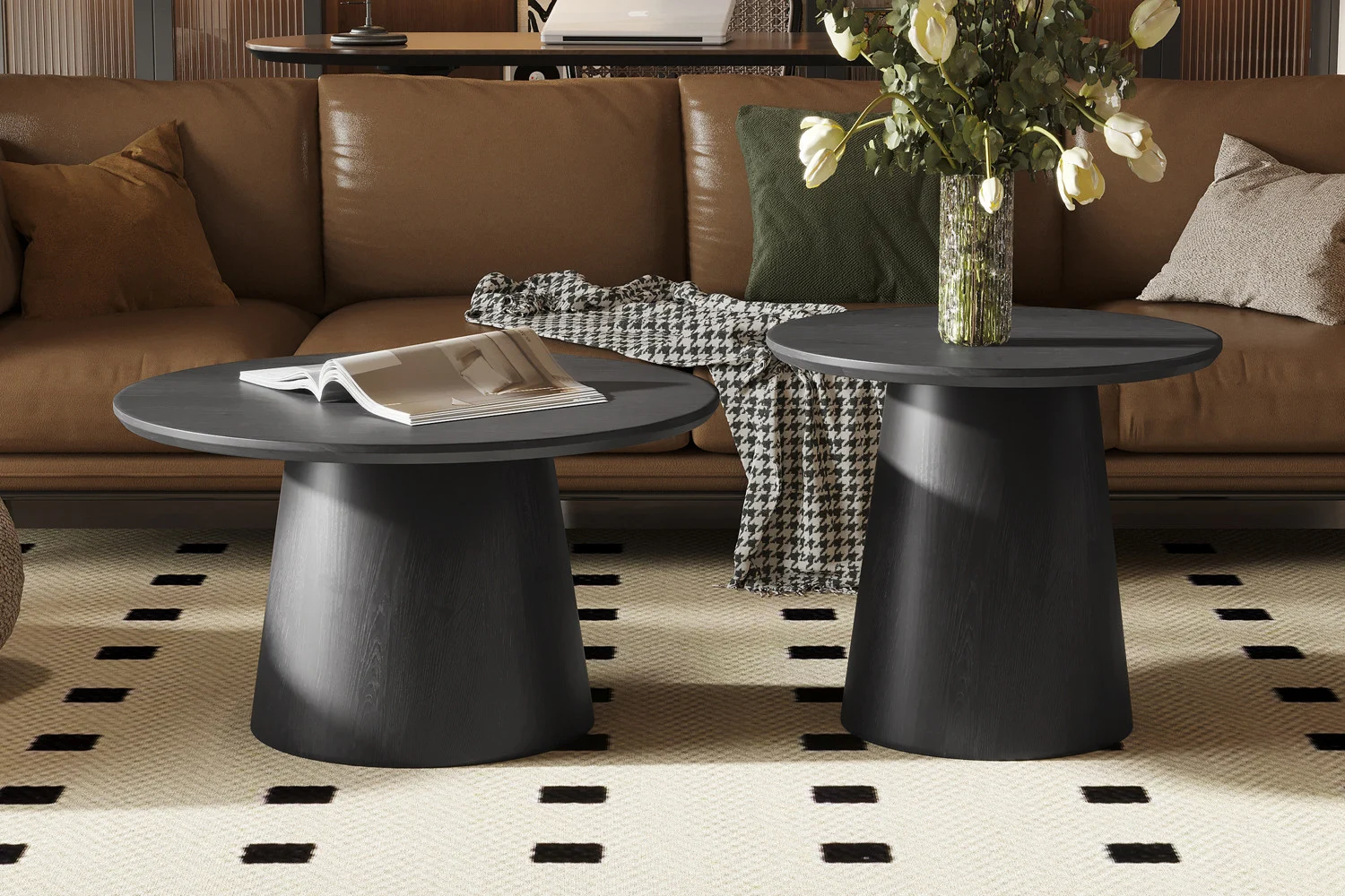 De Salontafel 80 CM Rond Design, in zwart, staat samen met een andere ronde salontafel op een vloerkleed met patroon in een moderne woonkamer. Op de ene ligt een tijdschrift, op de andere een vaas met bloemen. Daarachter staat een bruine bank met kussens.